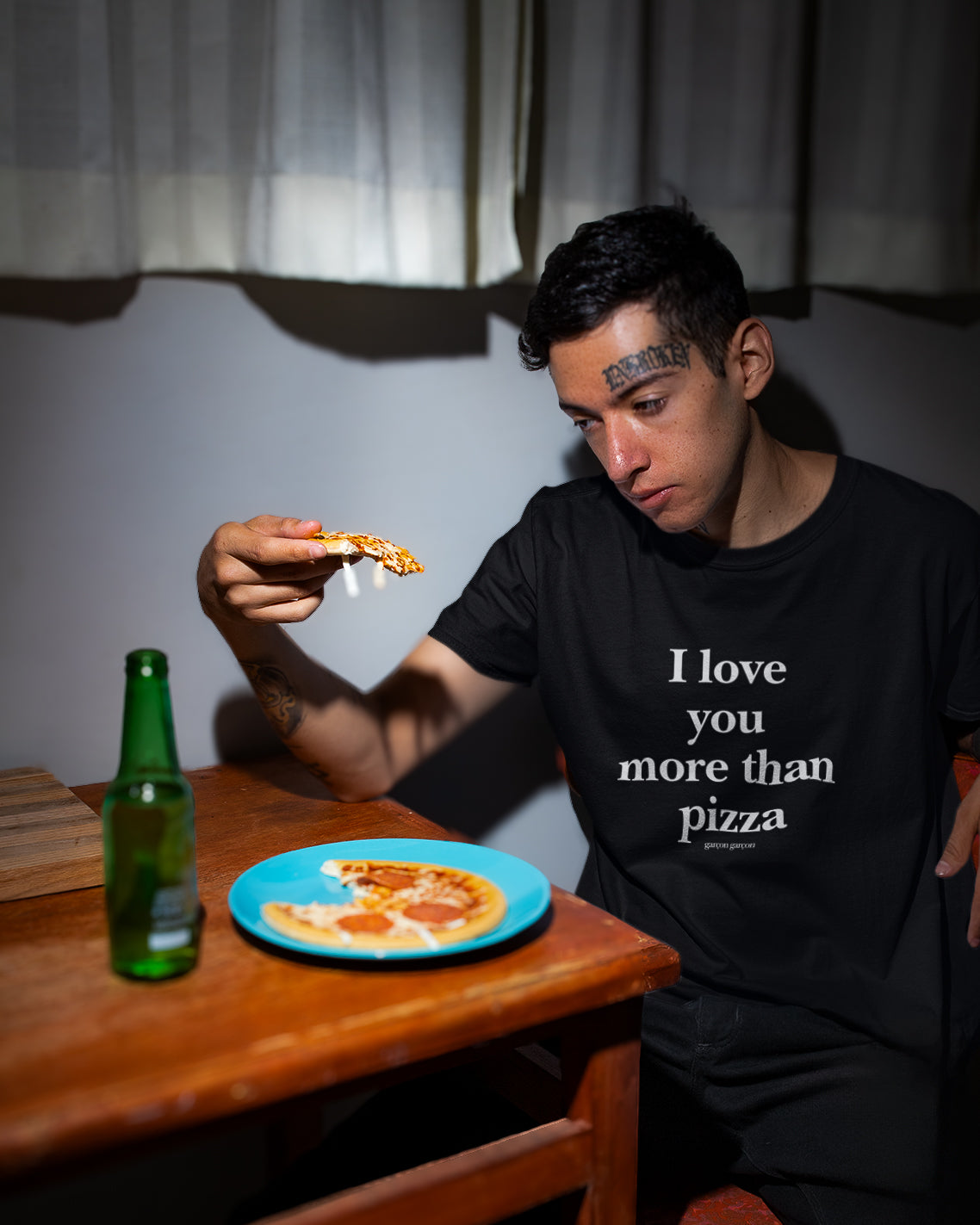 I love you more than pizza oversized t-shirt. A person seated at a wooden table, wearing a black t-shirt with the phrase "I love you more than pizza" written in white. They are glancing down, thoughtfully, at a slice of pizza in their hand. A partially eaten pizza sits on a blue plate, and a green bottle is to the side, against a backdrop of a dimly lit room with a window dressed in white curtains.