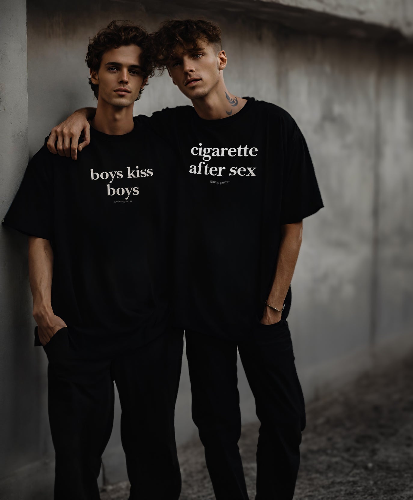 Two models stand side by side against a gray concrete wall, each sporting a bold statement t-shirt from Garçon Garçon. One t-shirt reads "boys kiss boys" and the other "cigarette after sex," both reflecting the brand's edge and commitment to free expression. They exude confidence and a cool, urban style that's signature to the Garçon Garçon aesthetic.