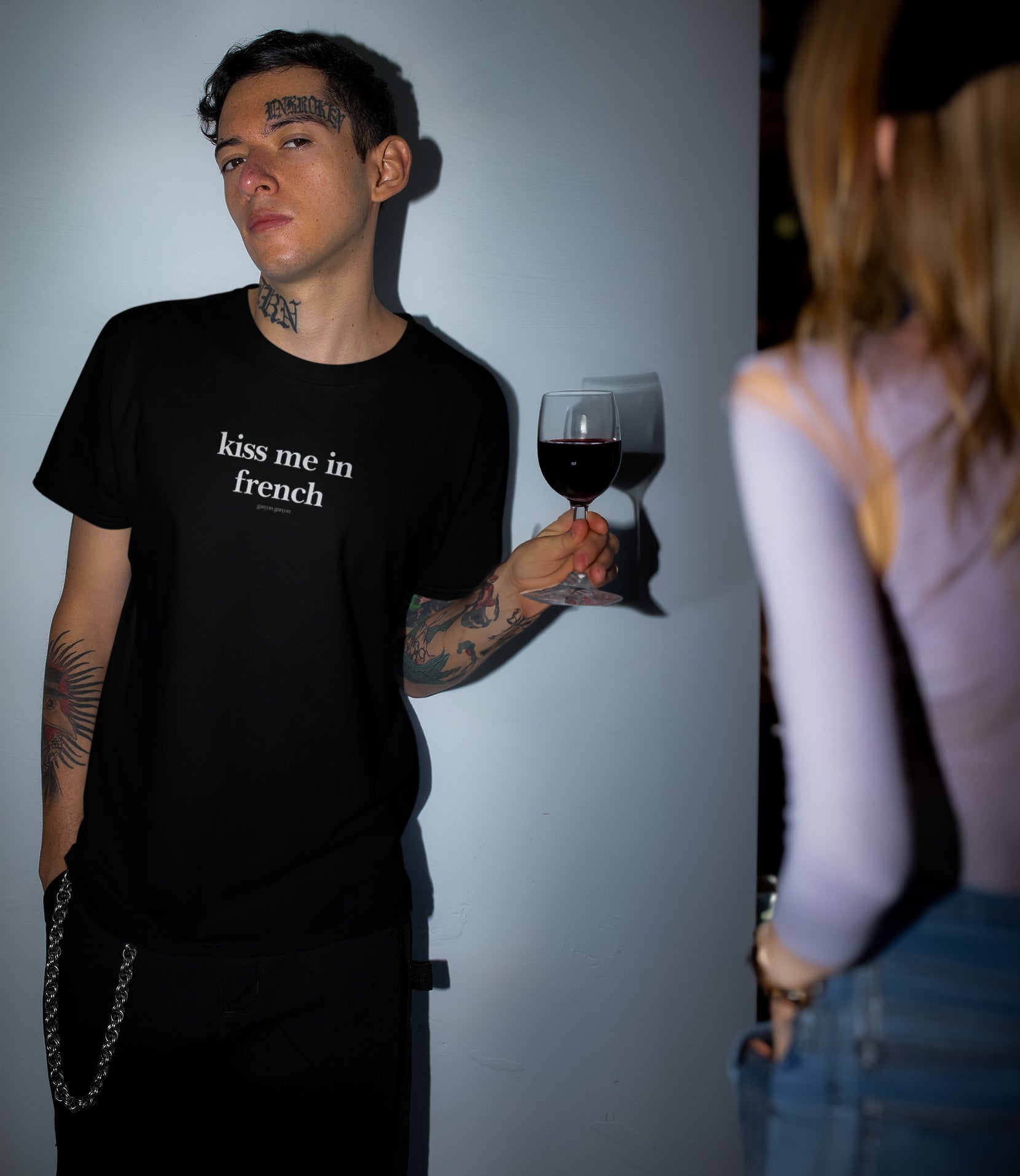 A man gazes thoughtfully away, a glass of red wine in hand, wearing a black t-shirt emblazoned with "kiss me in french" in crisp white lettering. The intimate moment is captured with soft lighting against a simple backdrop, highlighting the shirt's message and the urban, tattooed style of the wearer.